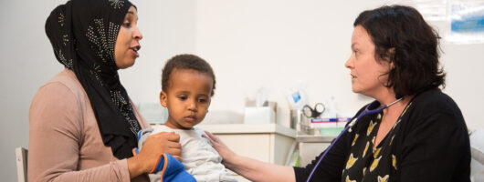 A photograph of young person with their child in a doctor's appointment. The child is sitting on their parents lap. The doctor is in conversation with the parent. The doctor is wearing a stethescope.