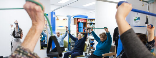 A photograph of older people in an exercise class. Everyone is seated with their arms raised above their head.