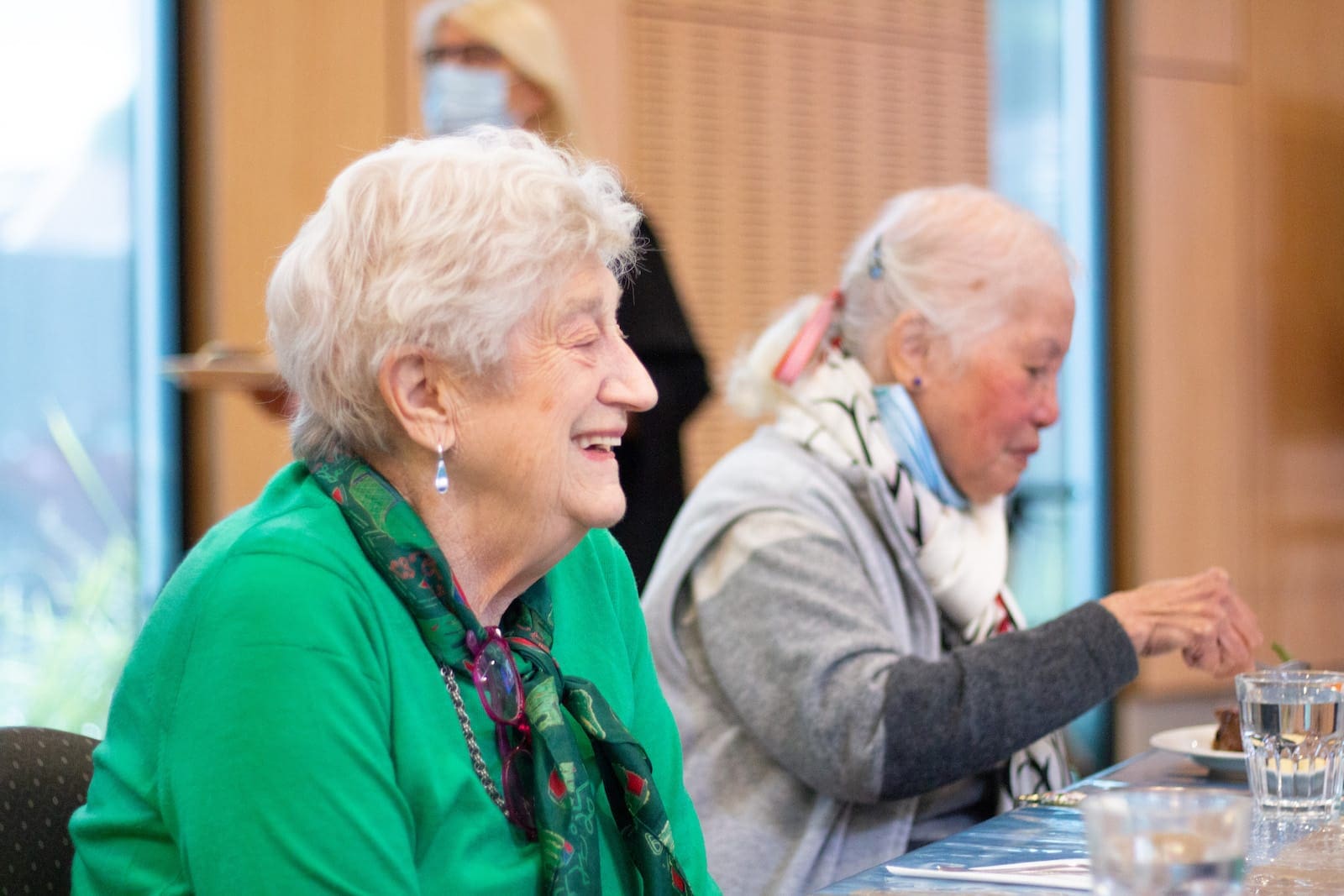 Several members of the community attending a session hosted by Older Persons Social Support and Respite. One is wearing a green shirt and scarf, the other is wearing a grey jumper.