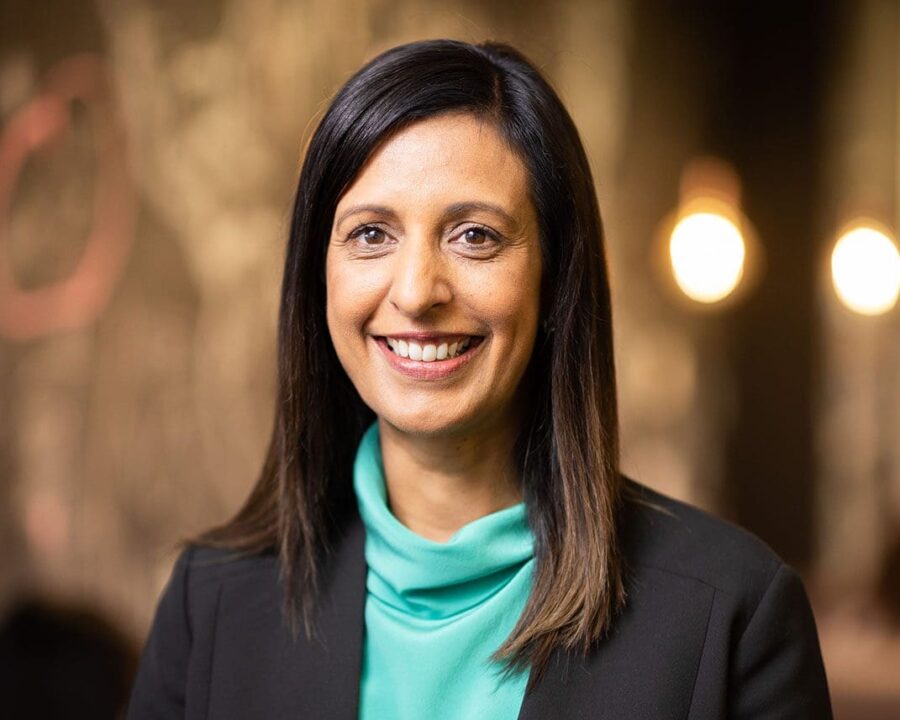 Nicole Bartholomeusz, cohealth's Chief Executive, wearing a dark grey jacket and a light blue shirt in front of a brown background.